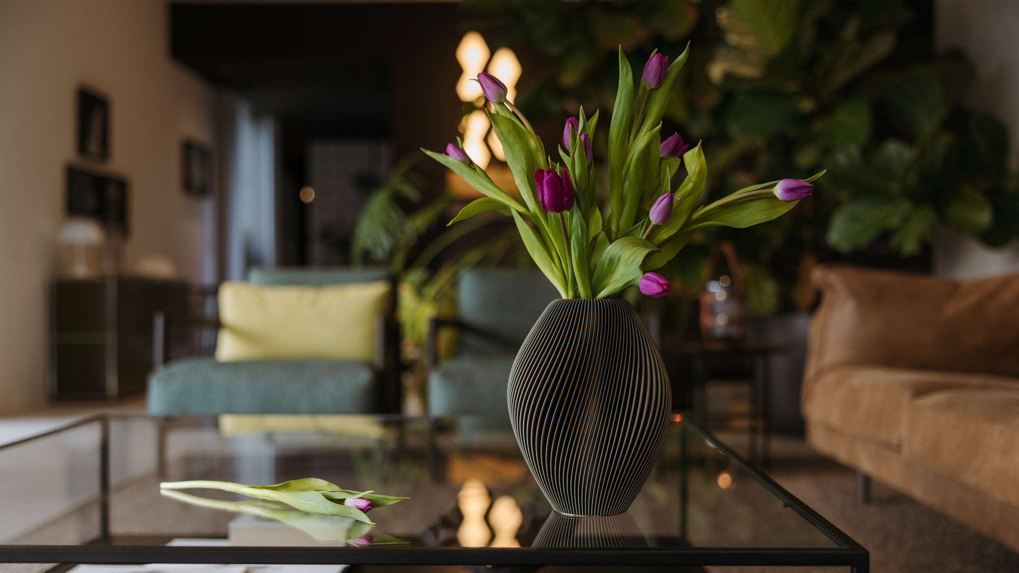 A dark green ribbed vase filled with purple tulips sits on a glass coffee table in a warmly lit living room, with sofas and plants softly blurred in the background.